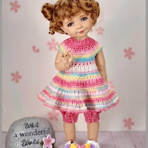 May include: A doll with curly brown hair styled in two buns, wearing a sleeveless, multi-colored, horizontally striped knit dress in shades of pink, yellow, blue, and white, paired with pink polka-dot bloomers and white shoes with floral accents.