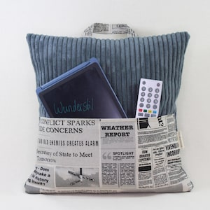 Könnte beinhalten: Ein blaues Cordkissen mit einer Tasche auf der Vorderseite. Die Tasche enthält ein Tablet und eine Fernbedienung. Das Kissen liegt auf einer Zeitung mit dem Aufdruck "Weather Report".