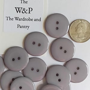 Peut inclure: Une collection de boutons ronds, lisses et gris clair à deux trous, disposés près d'une pièce de monnaie américaine et d'une carte blanche portant le texte The W&P The Wardrobe and Pantry. Les boutons ont une finition brillante.