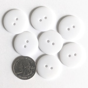 May include: A collection of seven white, round buttons with two holes each, arranged on a white surface. A U.S. quarter coin is also present, providing a size reference. The buttons are evenly spaced and appear to be made of a smooth material.