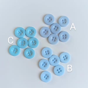May include: A selection of light blue buttons in varying shades, grouped together. Each button features four holes. The buttons are circular with a subtly raised rim. The background is a plain, light-coloured surface.