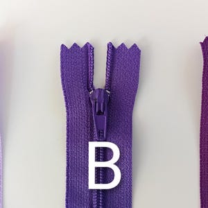 May include: Three zippers in shades of purple, labeled A, B, and C. The zippers are closed, showing the teeth and pull tabs. The background is a plain white surface. The zippers are for sewing and crafting.