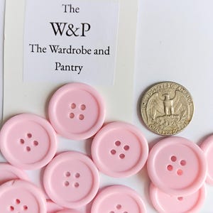 Puede incluir: Un conjunto de botones redondos de color rosa claro con cuatro agujeros cada uno. Una moneda está al lado de los botones para comparar el tamaño. El texto "The W&P The Wardrobe and Pantry" está en una tarjeta blanca.