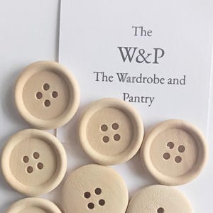 Puede incluir: Una colección de botones de madera de color beige claro, cada uno con cuatro agujeros centrales y un borde circular elevado. Una tarjeta blanca se sitúa detrás de los botones, mostrando el texto impreso: The W&P The Wardrobe and Pantry.