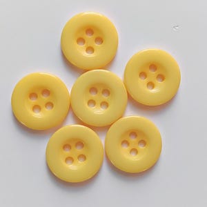 Może przedstawiać: Sześć okrągłych, żółtych guzików z czterema otworami każdy, ułożonych na białej powierzchni. Guziki mają błyszczące wykończenie i są równomiernie rozmieszczone. Te guziki nadają się do projektów krawieckich.