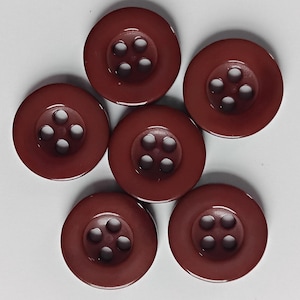 Puede incluir: Seis botones redondos de color rojo oscuro con cuatro agujeros cada uno están dispuestos sobre una superficie blanca. Los botones tienen un centro ligeramente elevado y un acabado brillante.