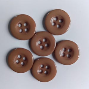 Puede incluir: Seis botones redondos marrones, cada uno con cuatro agujeros. Los botones son de un color uniforme y parecen estar hechos de plástico o un material similar. Están dispuestos en un grupo suelto.
