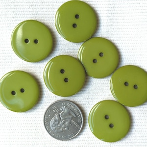 May include: A collection of eight olive green, round buttons with two holes each. A U.S. quarter is included for size comparison. The buttons have a glossy finish and are arranged on a white surface. These buttons are suitable for sewing and craft projects.