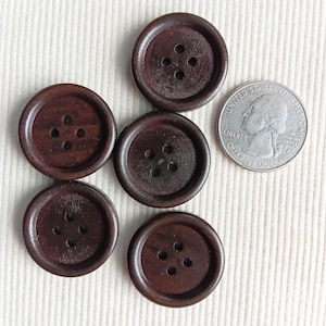 Puede incluir: Se muestran seis botones de madera marrón oscuro, cada uno con cuatro agujeros. Se incluye una moneda para comparar el tamaño. Los botones son redondos con un borde ligeramente elevado y un acabado liso. Están dispuestos sobre una superficie texturizada de color crema.
