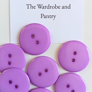 May include: A collection of round, smooth, lavender-colored buttons, each featuring two central holes. Positioned above the buttons is a white card with the printed text: The W&P The Wardrobe and Pantry. The buttons are arranged in a cluster.