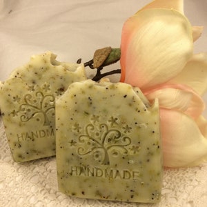 May include: Two handmade bars of soap with a green and white swirl design. The soap has a tree design with the word "HANDMADE" stamped on it. The soap is sitting on a white lace surface with a pink flower behind it.