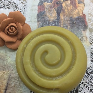 Puede incluir: Una pastilla de jab&oacute;n redonda de color verde con un dise&ntilde;o en espiral. El jab&oacute;n est&aacute; sobre un fondo de encaje blanco.