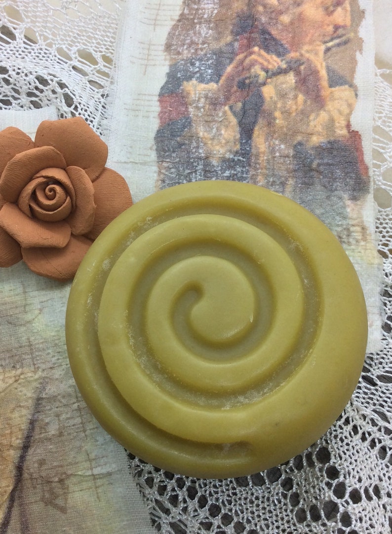 Puede incluir: Una pastilla de jab&oacute;n redonda de color verde con un dise&ntilde;o en espiral. El jab&oacute;n est&aacute; sobre un fondo de encaje blanco.