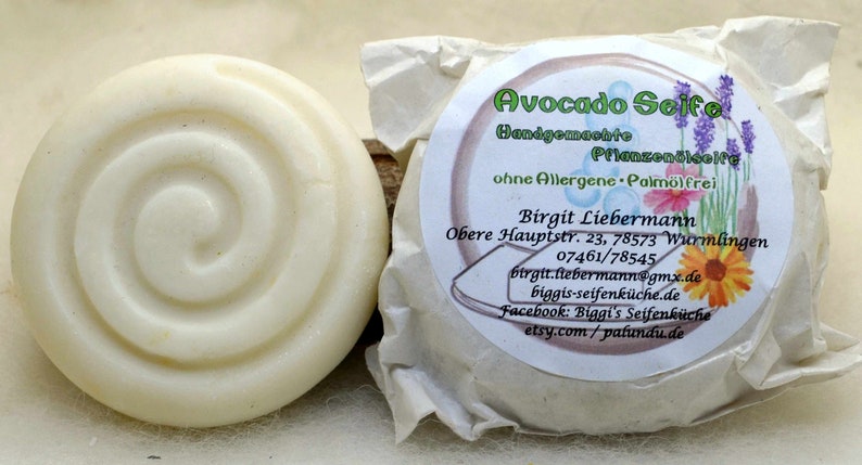 Puede incluir: Dos barras redondas de jab&oacute;n blanco, una envuelta en papel marr&oacute;n con una etiqueta que dice "Avocado Seife Handgemachte Pflanzen&ouml;le ohne Allergene - Palm&ouml;lfrei Birgit Liebermann Obere Hauptstr. 23, 78573 Wurmlingen 07461/78545 btrgit.ftebermann@gmx.de biggis-seifenk&uuml;che.de Facebook: Biggi's Seifenk&uuml;che etsy.com/pafundu.de".