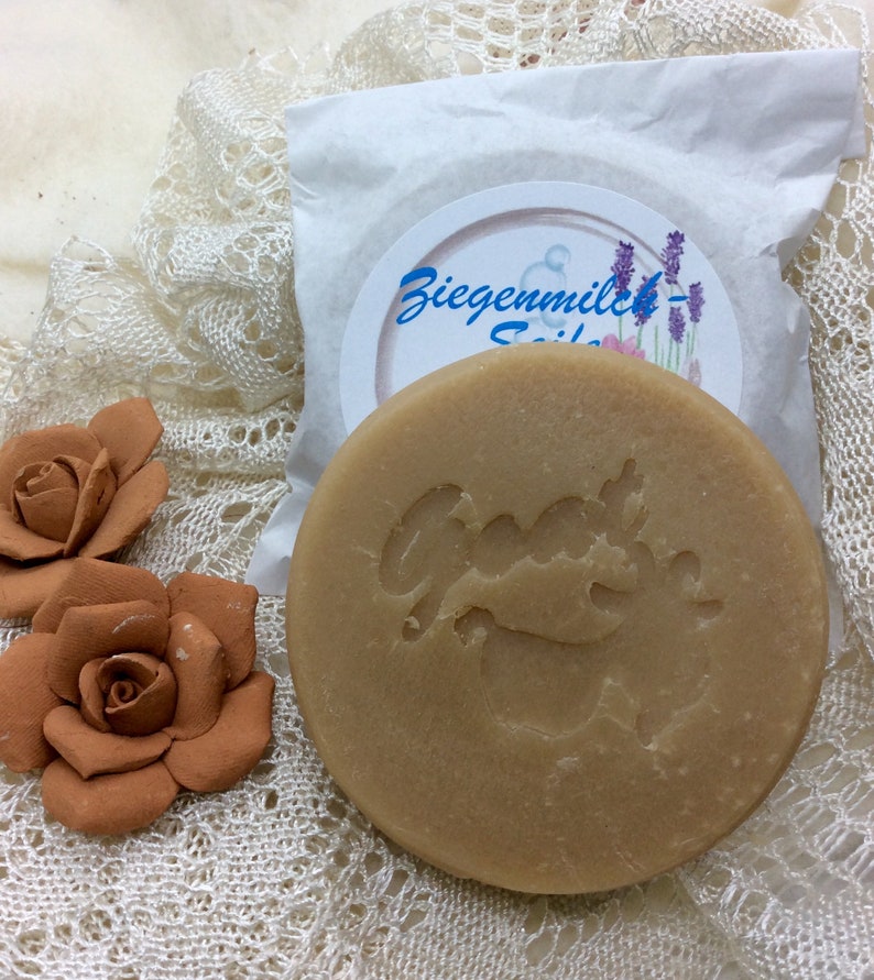 May include: A round, light brown bar of goat milk soap with the words "Ziegenmilch-Seife" and a goat logo on the label. The soap is sitting on a white lace surface.