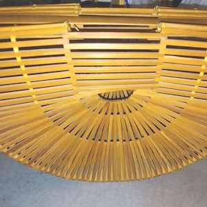 May include: A handcrafted, semi-circular bamboo handbag with a natural, light brown finish. The bag features a unique design with horizontal slats and a rounded, fan-like bottom. The top has a hinged closure. This stylish bag is perfect for carrying essentials.