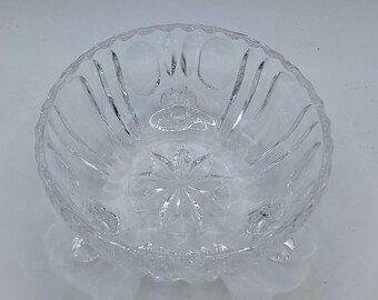 May include: Clear glass bowl with a starburst design in the center and a decorative rim. The bowl has a crown design and is supported by three small feet. The bowl is likely used for serving or display.