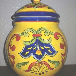 May include: A yellow ceramic jar with a blue lid and intricate floral designs in blue, red, and green. The jar has a round body and a wide mouth.