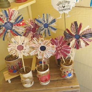 May include: A collection of six red, white, and blue fabric flowers in recycled cans. The flowers are made from repurposed fabric scraps and are arranged in a rustic farmhouse style. The sign above reads "Welcome to our Farmhouse".