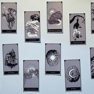 May include: A collection of tarot cards with grayscale illustrations. Each card features unique artwork and titles such as "The Fool," "The Hermit," and "The Devil." The cards are arranged on a white surface, showcasing a gothic or mystical theme.