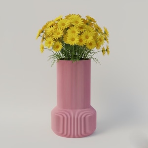 Puede incluir: Un jarrón rosa, con líneas verticales, contiene un vibrante ramo de margaritas amarillas. El jarrón tiene forma cilíndrica con una base y parte superior más anchas. Las margaritas están en plena floración, creando un contraste alegre con el jarrón rosa.