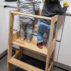 Può includere: Due bambini stanno in piedi su uno sgabello di legno, raggiungendo un bancone da cucina. Lo sgabello ha due gradini e un corrimano. I bambini indossano abiti casual.