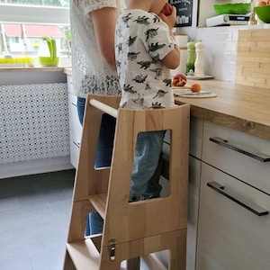 Peut inclure: Un tabouret d'apprentissage en bois à deux marches avec un loquet de sécurité. Un jeune enfant se tient sur la marche supérieure, atteignant un comptoir. L'enfant porte une chemise blanche et bleue à motif de dinosaure. Le tabouret est dans un environnement de cuisine.
