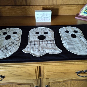 May include: A black Halloween table runner featuring three ghost-shaped appliqués. Each ghost has a black circular mouth and eyes, with patterned fabric details in cream and black. The text on a small wooden block reads, "I'm not Retired, I'm a PROFESSIONAL GRANDMA."