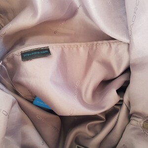 Puede incluir: El interior de una bolsa de color rosa claro con una etiqueta negra que dice "piquadro.com". La bolsa est&aacute; forrada con un tejido suave de color rosa claro y tiene un cierre de cremallera.