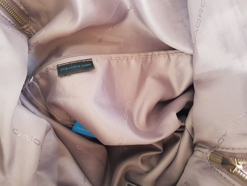 Puede incluir: El interior de una bolsa de color rosa claro con una etiqueta negra que dice "piquadro.com". La bolsa est&aacute; forrada con un tejido suave de color rosa claro y tiene un cierre de cremallera.