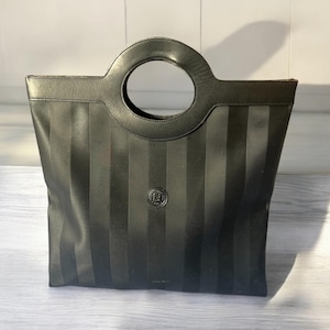 May include: Black tote bag with vertical stripes and a circular handle. The bag features a small circular emblem on the front. The handle is a large, circular design. The bag is made of a smooth material.