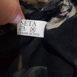 May include: A white tag with black text that reads "SETA" and "Made in Italy". The tag has a symbol of an iron and a circle with a slash through it. The tag is attached to a black and white patterned fabric.