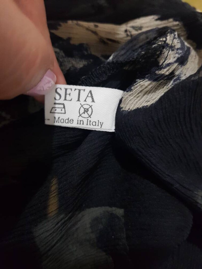 May include: A white tag with black text that reads "SETA" and "Made in Italy". The tag has a symbol of an iron and a circle with a slash through it. The tag is attached to a black and white patterned fabric.