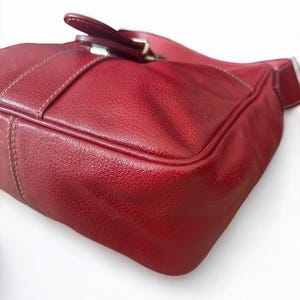 May include: A red leather handbag with a textured surface. The bag features a buckle closure and a shoulder strap. The stitching is visible in a lighter color, adding detail to the design. The bag is a deep shade of red.