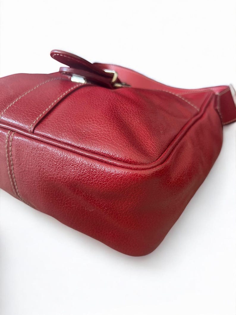 May include: A red leather handbag with a textured surface. The bag features a buckle closure and a shoulder strap. The stitching is visible in a lighter color, adding detail to the design. The bag is a deep shade of red.