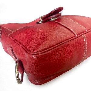 May include: A red leather handbag with white stitching and a silver-tone metal ring. The bag features a flap closure with a matching leather tab and a silver buckle. The leather has a textured appearance.