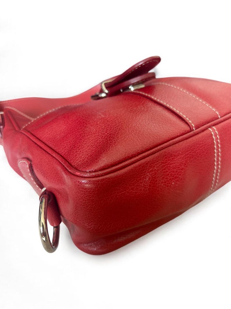 May include: A red leather handbag with white stitching and a silver-tone metal ring. The bag features a flap closure with a matching leather tab and a silver buckle. The leather has a textured appearance.