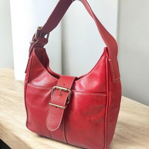 May include: A red leather shoulder bag with a curved shape. The bag features a buckle closure with a gold-colored metal buckle and a single shoulder strap. White stitching accents the edges of the bag.