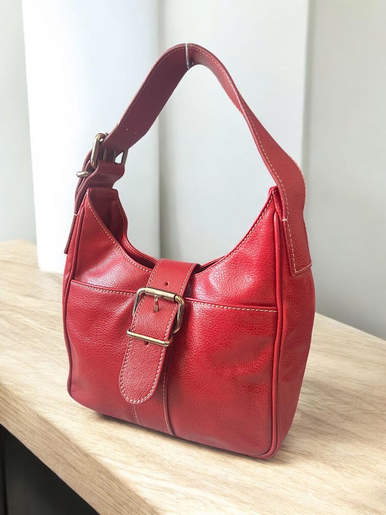 May include: A red leather shoulder bag with a curved shape. The bag features a buckle closure with a gold-colored metal buckle and a single shoulder strap. White stitching accents the edges of the bag.
