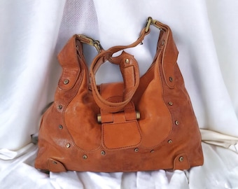 May include: A tan leather hobo bag with brass-coloured hardware. The bag has a curved shape, a top handle, and a flap closure. Small, round studs decorate the exterior. The bag is set against a white background.
