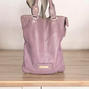 Puede incluir: Un bolso tote de cuero de color morado claro con herrajes dorados y cierre de cremallera.