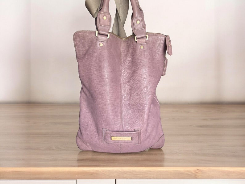 Puede incluir: Un bolso tote de cuero de color morado claro con herrajes dorados y cierre de cremallera.