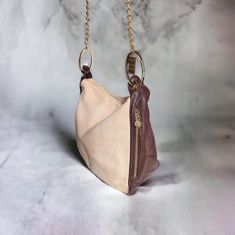 May include: A beige and brown leather shoulder bag with a gold chain strap and two gold rings. The bag has a zipper closure and a small gold tag.