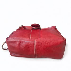 May include: A red leather handbag with a textured surface. The bag features a top handle, a silver-tone metal ring, and a buckle closure. White stitching accents the design.