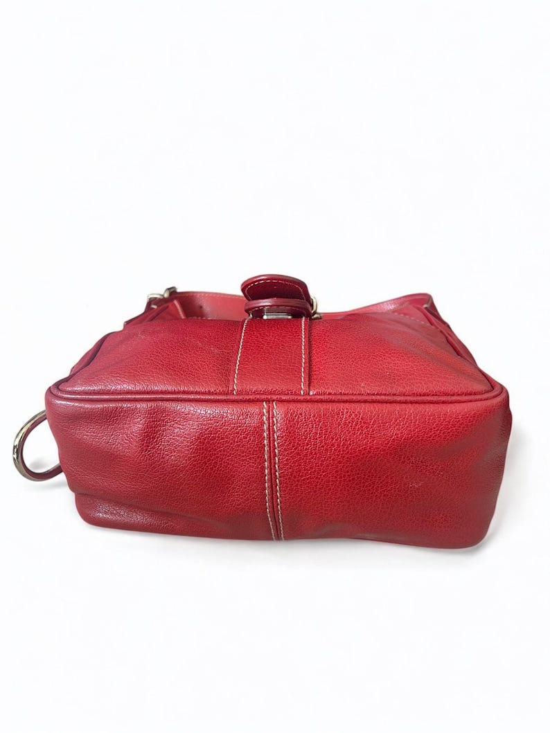 May include: A red leather handbag with a textured surface. The bag features a top handle, a silver-tone metal ring, and a buckle closure. White stitching accents the design.