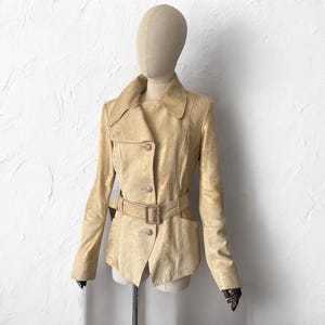 May include: A tan jacket with a textured finish, displayed on a mannequin. The jacket features a notched collar, button closure, and a matching belt with a rectangular buckle. The sleeves are long, and the jacket has a tailored fit.
