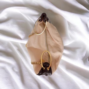 May include: A beige canvas handbag with brown leather accents and gold chain straps. The bag has two large gold rings on the front.