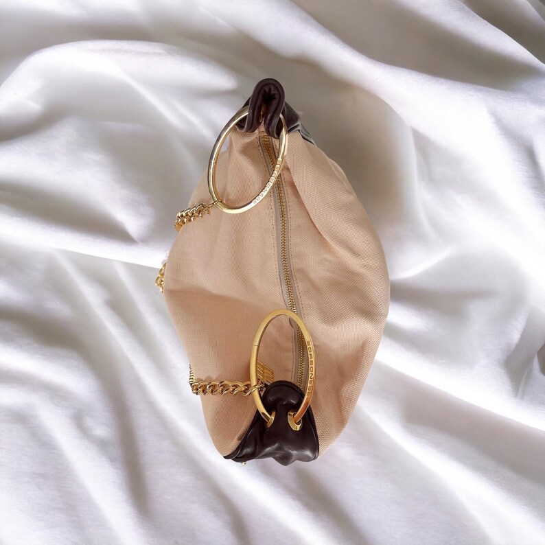 May include: A beige canvas handbag with brown leather accents and gold chain straps. The bag has two large gold rings on the front.
