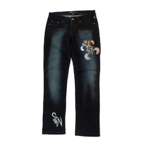 Könnte beinhalten: Dunkelblaue Jeans aus Denim mit silberner Stickerei eines stilisierten Blumenmusters auf dem rechten Bein und einem kleineren Muster auf dem linken Bein.