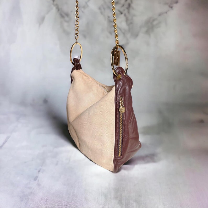 May include: A beige and brown leather handbag with a gold chain strap and a gold ring detail. The bag has a zipper closure and a flap.